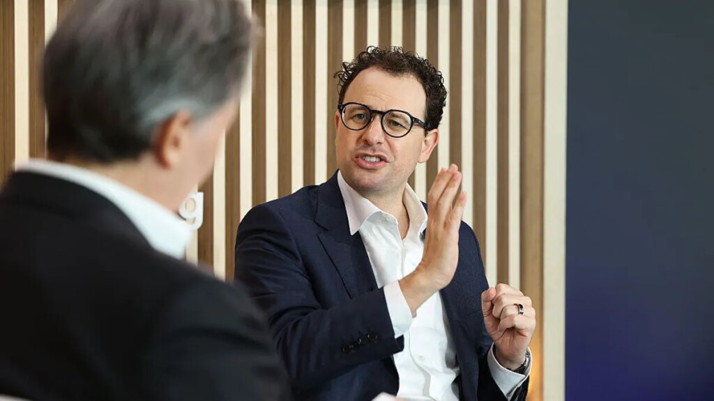Trump Slams Anthropic as ‘Woke,’ Orders Feds to Stop Using Claude AI Anthropic CEO Dario Amodei speaks while seated at a conference forum, with the back of another person's head out of focus in the foreground.
