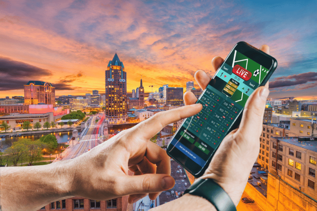 Wisconsin mobile sports betting bill heads to Senate New Wisconsin Assembly advances Tribal-controlled statewide mobile sports betting plan. Close-up of a person holding a smartphone displaying a live sports betting app, with a city skyline at sunset in the background, featuring high-rise buildings and a river running through downtown.