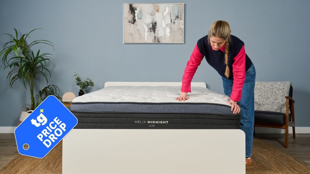 A light haired mattress tester wearing a pink shirt and blue jeans stands next to a blue and white mattress, placing her hands on its cover as she inspects it.