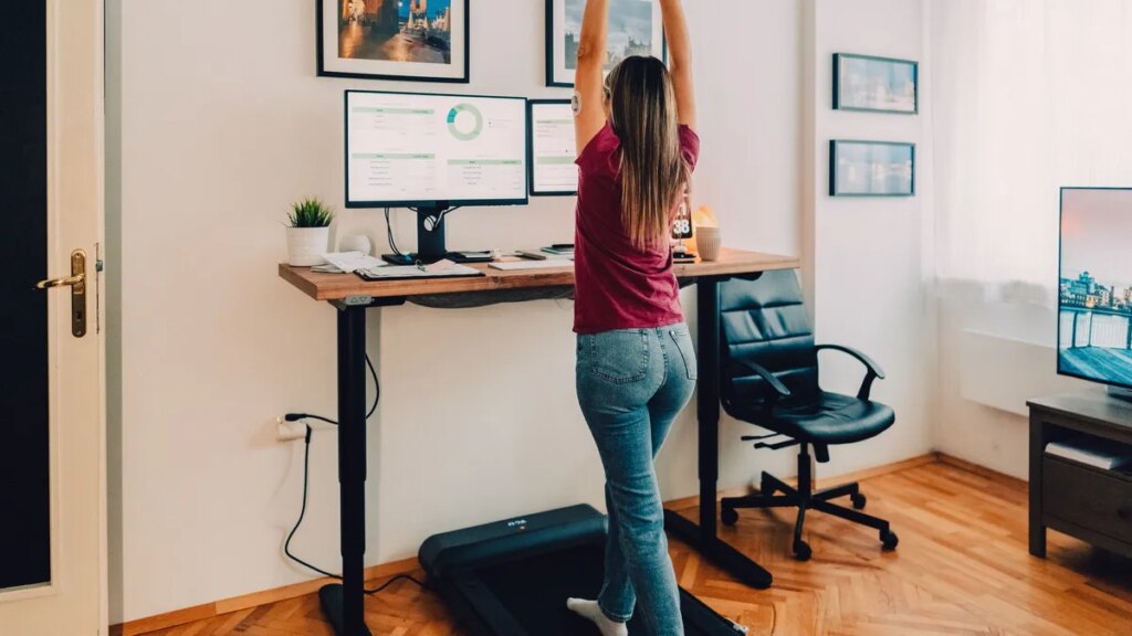 The Wellness Devices Fitness Professionals Actually Use and Swear By A person in jeans and a dark pink-purple tee using an under-desk treadmill while working on the computer.