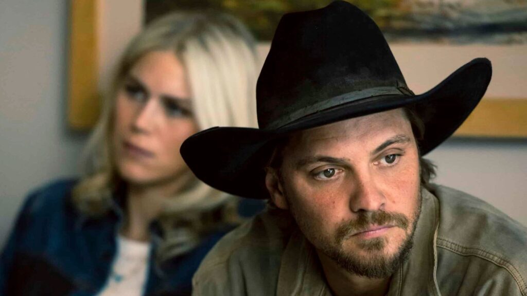 Kayce looks off into the distance while a blonde woman is sat behind him