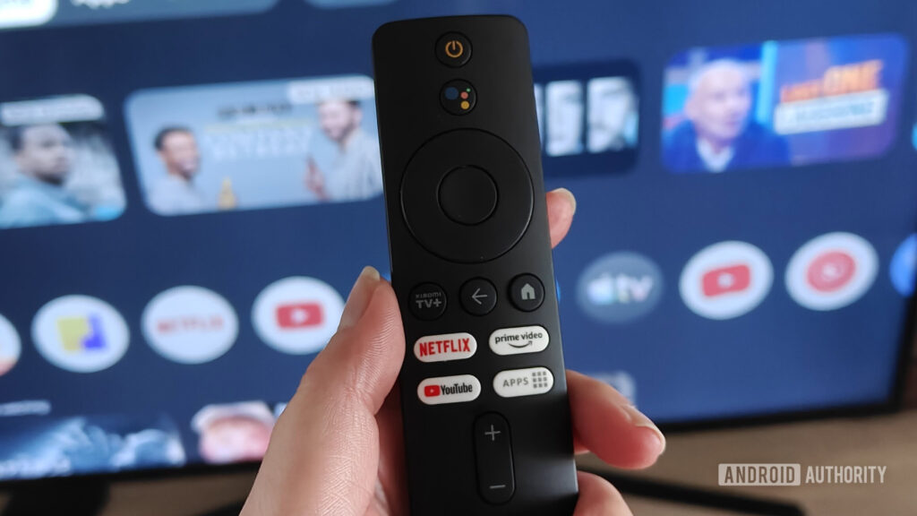 a remote held in a hand with the google tv interface on a screen in the background