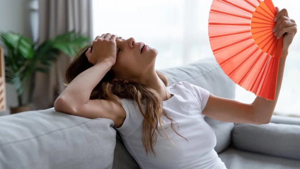 Summer Blackout To-Do List: 9 Things to Keep Everyone Cool and Safe An overheated woman with her hand on her forehead, fanning herself with a red hand fan, on a light gray couch.